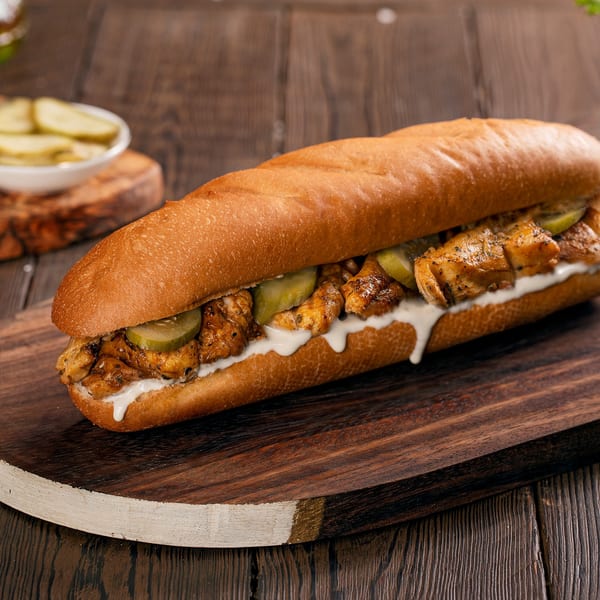 Grilled chicken sub sandwich with pickles and creamy sauce on a rustic wooden board.