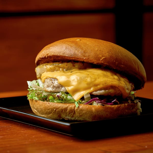 Gourmet burger with melted cheese, grilled pineapple, lettuce, and red cabbage on a black plate.