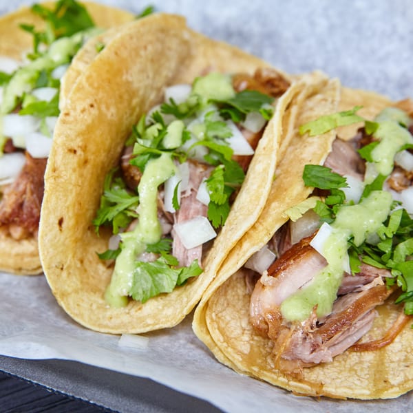 Three carnitas tacos with chopped onions, cilantro, and green salsa, served on parchment paper.
