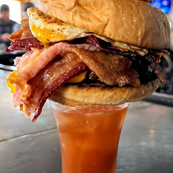 Massive burger with bacon and fried egg balanced on a Bloody Mary, garnished with a fried chicken strip.